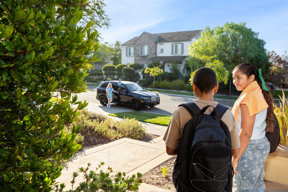 Two backpacked youths at a home entrance; branded black vehicle approaching.