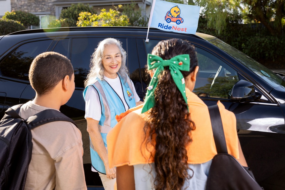 Uniformed older adult greeting two youths beside a branded black vehicle.